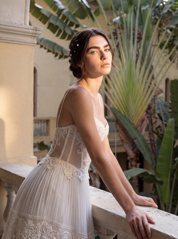 A model in a pleated white gown with lace trim, leaning on a stone balcony. AI-generated image.
